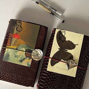 May include: Two brown leather journals with decorative elements. One has a vintage art print and a mother-of-pearl button, the other a butterfly illustration and a silver heart charm. Two pens are also visible.