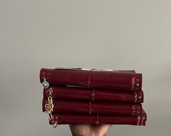 May include: A stack of four burgundy-colored journals held in a hand. Each journal has a decorative charm attached to the side. The journals are bound with a red elastic band.