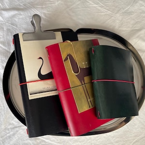 Könnte beinhalten: Drei ledergebundene Notizbücher in Schwarz, Rot und Dunkelgrün. Das schwarze Notizbuch hat eine silberne Metallklammer und eine Schwanenillustration mit dem Text SELBY HOUSE. Das rote Notizbuch enthält eine goldfarbene Messingklammer und ein Pferdemotiv.