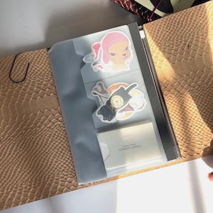 Peut inclure: Carnet ouvert avec une couverture marron texturée et une poche transparente contenant des autocollants. Les autocollants représentent des personnages et des objets de dessins animés. Une carte de visite est visible dans la poche. Le carnet est ouvert sur une surface blanche.