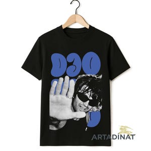 以下が含まれることがあります： サングラスをかけた人物と、顔の前に手をかざしたグラフィックが特徴の黒いTシャツ。「D3O」の文字が青色で画像の上にあります。木製のハンガーにTシャツがかけられています。