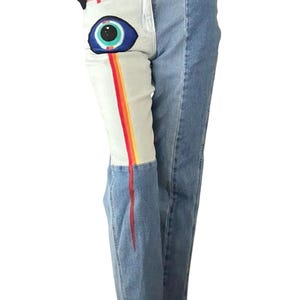 May include: Light wash denim jeans with a unique design. One leg features a large eye graphic with a rainbow stripe, while the other leg is plain. The jeans have a classic five-pocket design and a straight leg fit.