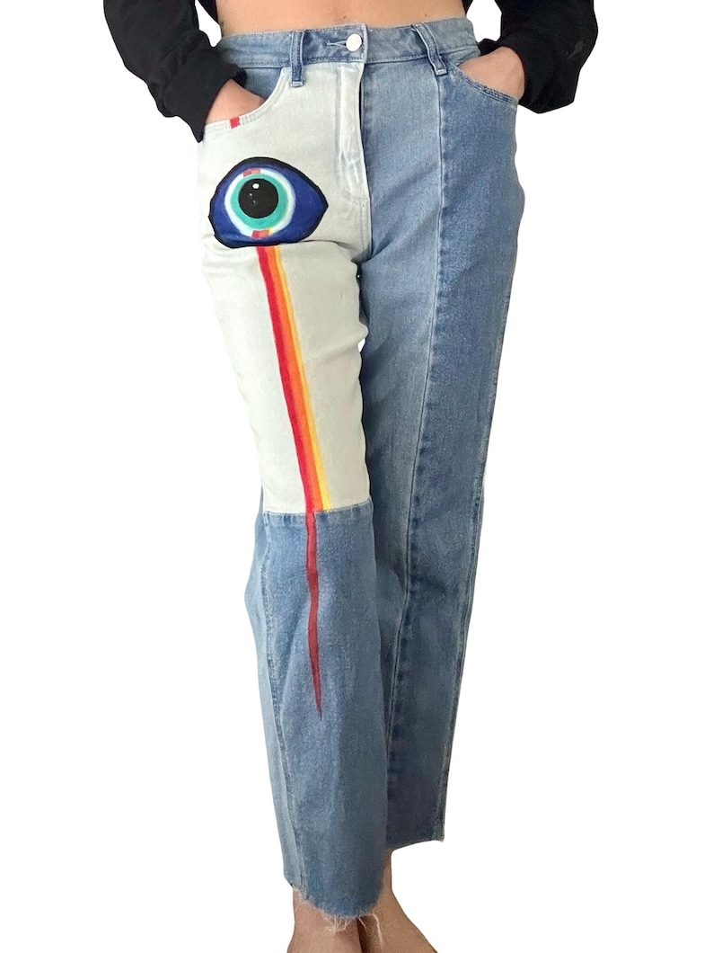 May include: Light wash denim jeans with a unique design. One leg features a large eye graphic with a rainbow stripe, while the other leg is plain. The jeans have a classic five-pocket design and a straight leg fit.