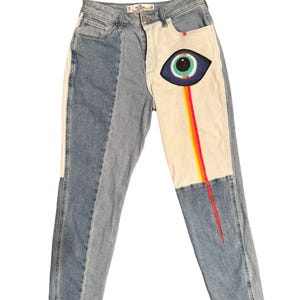 May include: Light wash denim jeans with a unique design. The jeans feature a two-tone color scheme with a white panel on the right leg. A painted eye graphic with a rainbow streak is on the white panel.