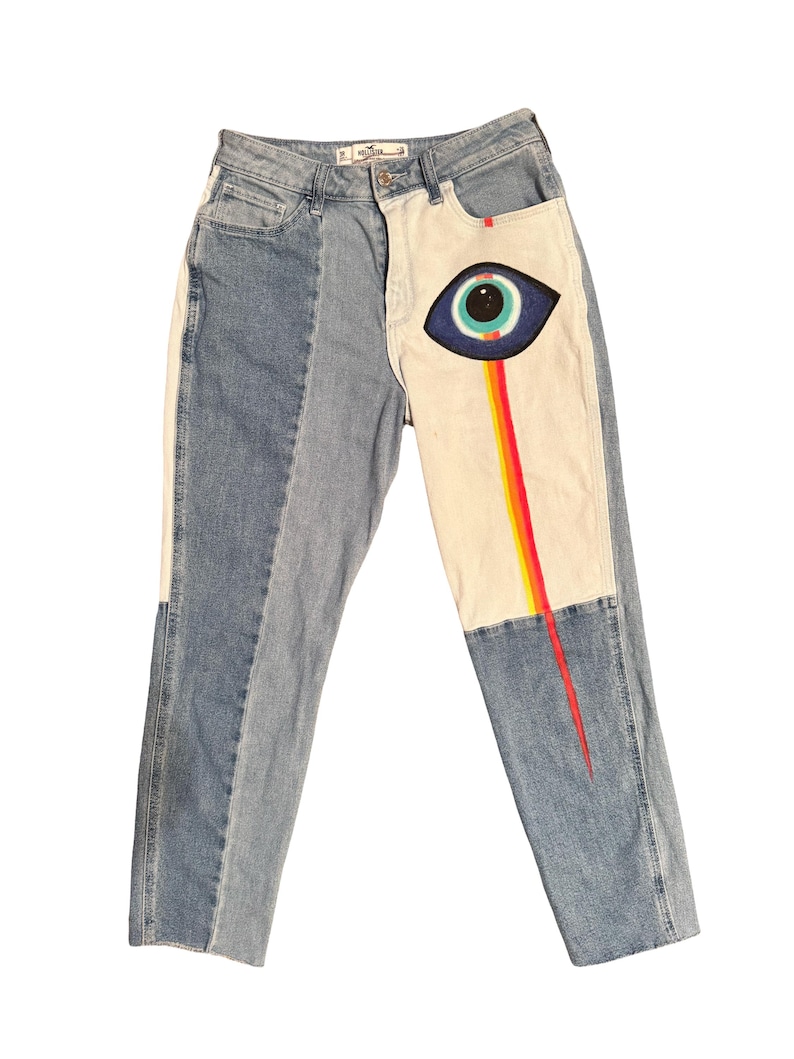 May include: Light wash denim jeans with a unique design. The jeans feature a two-tone color scheme with a white panel on the right leg. A painted eye graphic with a rainbow streak is on the white panel.