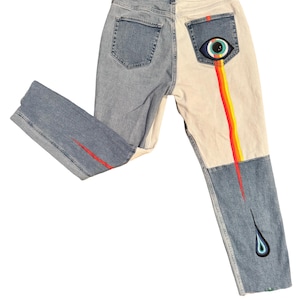 May include: Light wash denim jeans with a two-tone design. One side is light blue, the other is off-white. The back pocket features an eye design with a rainbow-colored stream. A red line accents the front of the left leg. The right leg has a teardrop design.