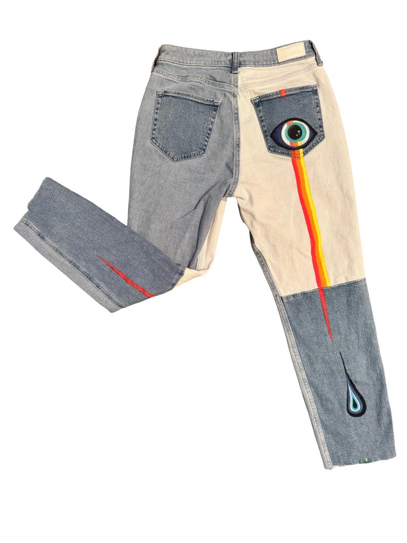 May include: Light wash denim jeans with a two-tone design. One side is light blue, the other is off-white. The back pocket features an eye design with a rainbow-colored stream. A red line accents the front of the left leg. The right leg has a teardrop design.