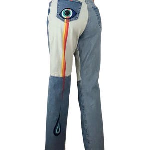 May include: Light wash denim jeans with a unique design. The back pocket features an eye graphic. A rainbow stripe runs down the left leg, with a teardrop design at the hem. The jeans have a straight leg cut.