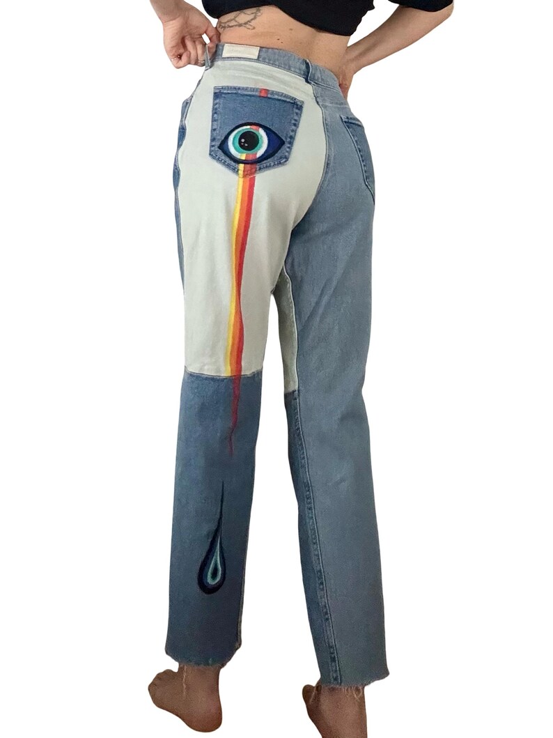 May include: Light wash denim jeans with a unique design. The back pocket features an eye graphic. A rainbow stripe runs down the left leg, with a teardrop design at the hem. The jeans have a straight leg cut.