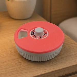Può includere: Un organizzatore di pillole settimanale color corallo e bianco. Il contenitore rotondo ha un coperchio rotante con i giorni della settimana etichettati. La base è bianca con scanalature verticali. Una piccola pianta in vaso bianco è sullo sfondo.
