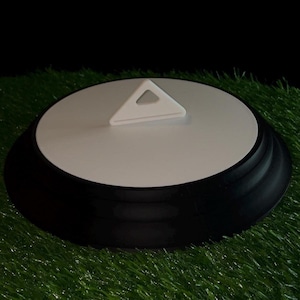 May include: A round, black and white object with a triangular handle. The object has a white top and a black base. The handle is white and has a triangular shape with a hole in the center. The object is sitting on a green surface.