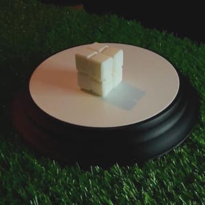 May include: A small, off-white cube-shaped object with a textured surface, resting on a white circular platform. The platform sits on a black base, with a green, artificial grass background.