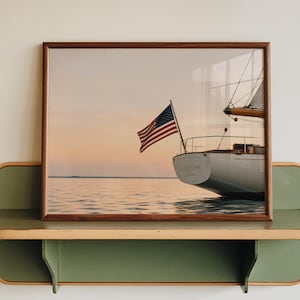 May include: A framed photograph of a sailboat stern with an American flag flying, set against a calm sunset over water. The print is housed in a dark wood frame and rests on a sage green wall-mounted shelf with light wood trim edges.