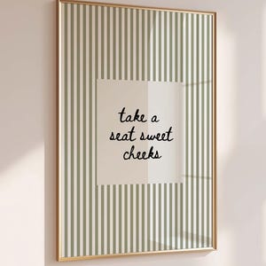 May include: A framed print with a gold-colored frame. The artwork features a striped background in shades of green and white. A square in the center displays the text "take a seat sweet cheeks" in a handwritten font. The print is displayed on a light-colored wall.