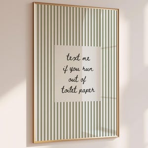 Puede incluir: Un marco dorado con una impresión de fondo a rayas verdes y blancas. Un cuadrado blanco en el centro contiene el texto: "text me if you run out of toilet paper."