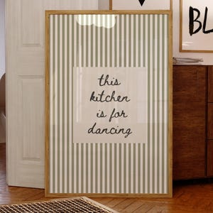 May include: A framed print with a light brown wooden frame. The print has a striped background in shades of green and white. The text "this kitchen is for dancing" is centered on the print. Two other framed prints are visible in the background.