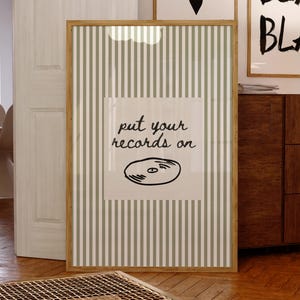 Op de afbeelding: Een ingelijste print met een lichtbruine houten lijst. De print heeft een gestreepte achtergrond met de woorden "put your records on" en een vinylplaat-illustratie. Twee andere ingelijste prints zijn zichtbaar op de achtergrond.