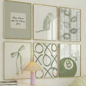 May include: A gallery wall featuring six framed art prints with gold frames. The prints include a leopard, lips, a bow, an 8-ball, and the text "slow down you're doing fine". A white cabinet with a lamp and a yellow chair complete the scene.