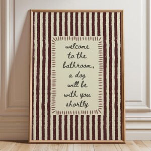 Può includere: Opera d'arte da parete incorniciata con sfondo color crema e strisce verticali bordeaux. L'opera presenta un riquadro rettangolare color crema con il testo "welcome to the bathroom, a dog will be with you shortly" in corsivo nero. La cornice è di colore marrone chiaro.