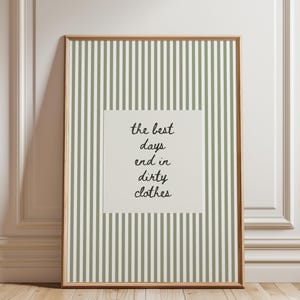 Peut inclure: Une affiche encadrée rectangulaire présentant un motif à rayures verticales vert sauge et blanc. Au centre, un carré blanc cassé contient le texte manuscrit noir : the best days end in dirty clothes, disposé sur quatre lignes.