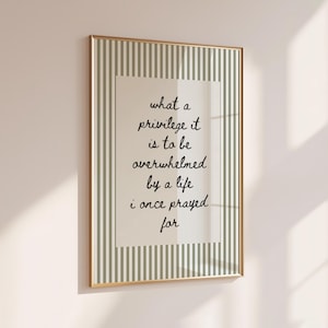 May include: A rectangular wall art print in a thin gold frame featuring a vertical sage green and white striped border surrounding a cream-colored center panel with the black handwritten text: what a privilege it is to be overwhelmed by a life i once prayed for.