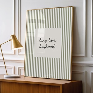 May include: A framed vertical wall art print featuring a sage green and white vertical striped pattern. In the center, a solid light beige rectangular block contains the black handwritten text that reads long live boyhood.