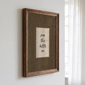 Può includere: Un quadro rettangolare con cornice in legno marrone scuro e sfondo marrone a righe verticali. Al centro, una toppa in tessuto beige con bordi dentellati riporta il testo scritto a mano in nero "pop the kettle on" su quattro righe.