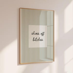 Op de afbeelding: Een rechthoekige wandprint in een dunne gouden lijst met een verticaal streeppatroon in saliegroen en wit. In het midden bevindt zich een wit vierkant met de zwarte cursieve tekst die luidt, shoes off bitches.