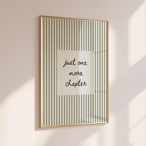 Pode incluir: Uma impressão emoldurada com uma moldura dourada. A impressão apresenta um fundo listrado em tons de verde e branco. Um quadrado central contém o texto manuscrito "just one more chapter" em preto.