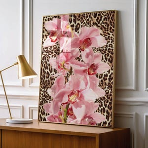 May include: A framed art print featuring pink orchids against a leopard print background. The orchids have red and yellow accents. The frame is gold, and the artwork is displayed on a wooden cabinet with a gold lamp.