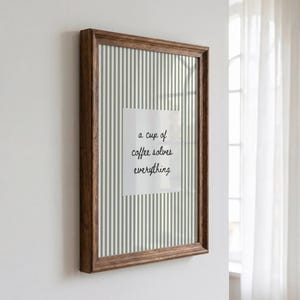 May include: A rectangular wall art piece in a dark brown wooden frame featuring a vertical sage green and white striped background with a central white square containing the black cursive text, a cup of coffee solves everything.