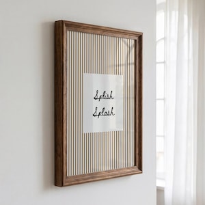 May include: A rectangular wall art piece in a dark brown wooden frame. The print features a white background with thin vertical gold stripes and a central white square containing the black cursive text Splish Splash centered in the middle.