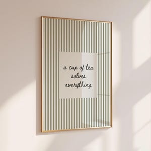 May include: A rectangular wall art print in a thin gold frame featuring a vertical sage green and white striped pattern. Centered on the print is a white square containing the black cursive text, a cup of tea solves everything.