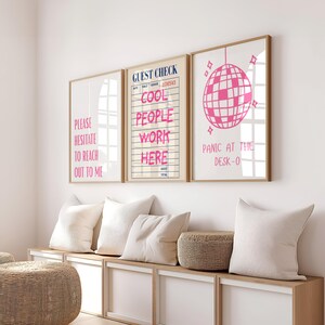 以下が含まれることがあります： 明るい木製フレームに入った3枚の壁掛けアート。左のプリントにはピンク色でPLEASE HESITATE TO REACH OUT TO MEと書かれています。中央はゲストチェックのフォームでCOOL PEOPLE WORK HEREとピンク色で書かれています。右のプリントにはピンク色のミラーボールが描かれています。