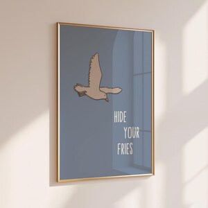 Op de afbeelding: Een ingelijste wandkunstprint met een lichtbruine meeuw die vliegt tegen een effen leiblauwe achtergrond. De tekst HIDE YOUR FRIES staat in witte hoofdletters in de rechterbenedenhoek van de print, gevat in een dunne gouden lijst.
