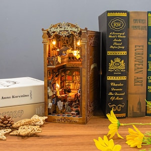 May include: A miniature wooden bookshelf diorama with the words "Magical Library" on top. The interior is lit with warm lights, showcasing a detailed library scene. Books and decorative elements are visible, alongside several books.