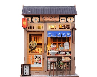 May include: A miniature wooden ramen shop model with a dark grey roof, a red lantern, and the Japanese text "もりたや". The shop features a detailed interior, including a counter, stools, and a cat figurine. The model is designed with intricate details.
