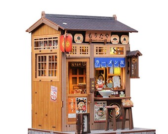 May include: A miniature wooden ramen shop model with a grey roof, wooden walls, and a brick base. The shop features a red lantern, signs with Japanese text, and a small interior with a ramen bowl. The model is lit from within.