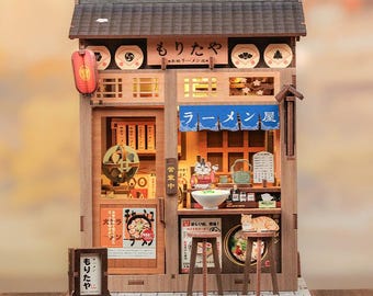 May include: A miniature wooden ramen shop model featuring a dark tiled roof, a blue fabric awning, and interior lighting. The facade displays Japanese text, a red lantern, and two stools. Inside, a cat figurine sits at a counter with a bowl of ramen.