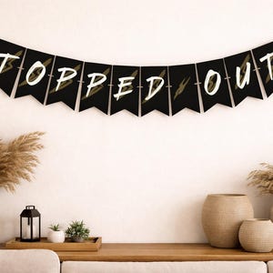 May include: A black banner with white text that reads "TOPPED OUT" is strung across the top of the image. The banner is decorated with gold lightning bolt designs. The banner is hanging above a wooden table with decorative vases and plants.