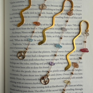 Op de afbeelding: Goudkleurige metalen bladwijzers in de vorm van een slang, versierd met kleurrijke edelstenen en kleine bedels. De bladwijzers liggen op de pagina's van een boek. De bedels bevatten dierenriemtekens.