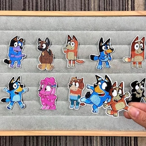 Könnte beinhalten: Eine Sammlung von aufgestickten Aufnähern mit Cartoon-Hund-Charakteren in verschiedenen Posen und Farben. Die Aufnäher werden auf einer grauen Oberfläche in einem Holzrahmen präsentiert. Die Hunde haben blaues, braunes und rosa Fell.