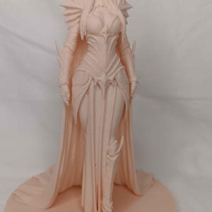 May include: A light peach-colored figurine of a female elf in a flowing gown and armor. The elf has long hair, pointed ears, and is standing on a round base. The figure is detailed with intricate designs.
