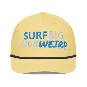Può includere: Un cappellino da baseball giallo pallido con una corda blu scuro alla base della corona. La parte anteriore presenta un testo ricamato in blu e bianco che recita SURF BIG RIDE WEIRD, con la parola woodlife in piccole lettere bianche.