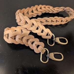 May include: Two tan leather purse straps with a braided design. Each strap has a brass-coloured clasp. One strap is coiled, and the other is straight.