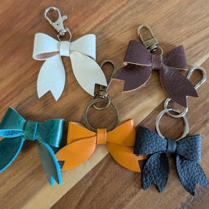 Könnte beinhalten: Eine Sammlung von Leder-Schlüsselanhängern in Form von Schleifen in verschiedenen Farben, darunter Weiß, Braun, Grün, Orange und Schwarz. Jede Schleife ist an einem Metallverschluss und Schlüsselring befestigt. Die Schlüsselanhänger liegen auf einer Holzoberfläche.