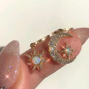 Op de afbeelding: Goudkleurige oorbellen met een hemels thema. Eén oorbel heeft een zonmotief met een opaalachtige steen, de andere een halve maan met een ster en opaalaccenten. De oorbellen zijn versierd met kleine, heldere kristallen.