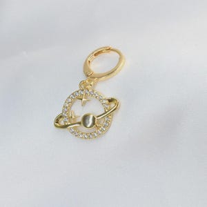 Op de afbeelding: Een goudkleurige oorbel met een planeetontwerp. De planeet is omcirkeld door een ring en een band van kleine, heldere kristallen. Twee sterren zijn ook in het ontwerp verwerkt. De oorbel heeft een scharnierende sluiting.