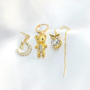 Op de afbeelding: Een verzameling van drie goudkleurige oorbellen. Eén heeft een maan- en sterontwerp, een andere een teddybeer en de derde een uil met een hangende ketting. Elke oorbel heeft een heldere, sprankelende steen.