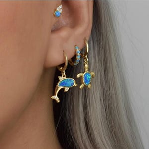 Op de afbeelding: Gouden oorbellen met dolfijn- en zeeschildpadhangers, beide met blauwe opaalachtige inleg. Een kleine oorbel met blauwe stenen is ook zichtbaar. De oorbellen worden op een oor gepresenteerd tegen een grijze achtergrond.